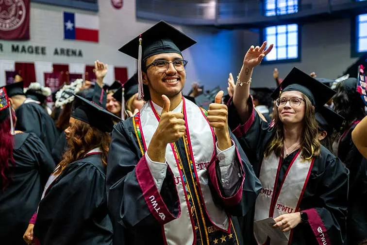 A man in academic regalia holds a thumbs up to the audience at commencement.