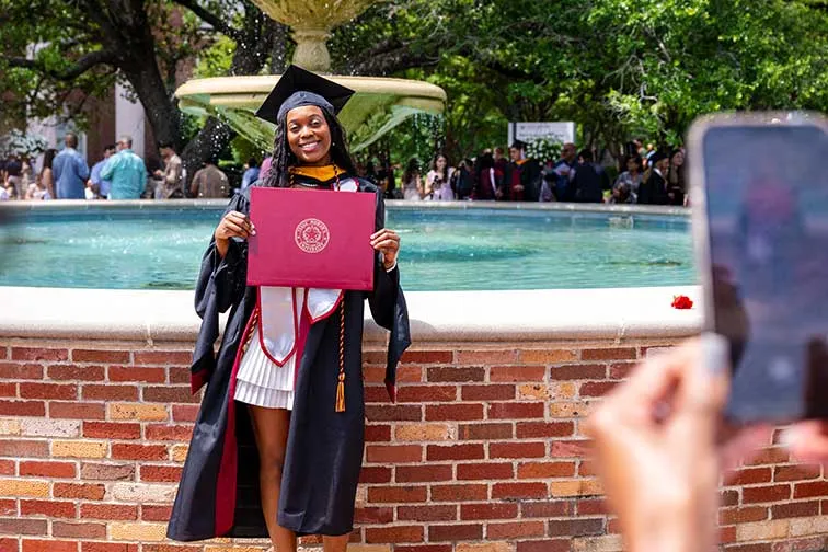 A woman holds her diploma holder from Commencement in front of the TWU fountain.