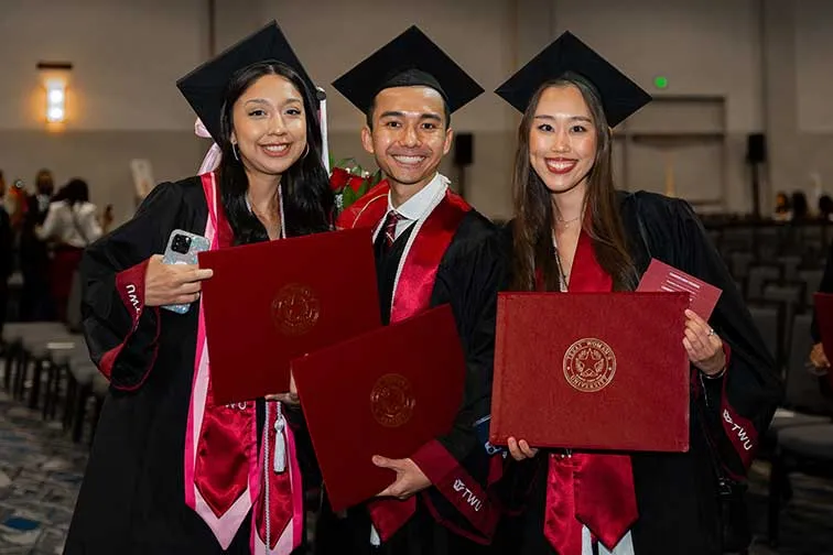 Two women and a man pose for a photo while in academic regalia and holding their diploma holders.