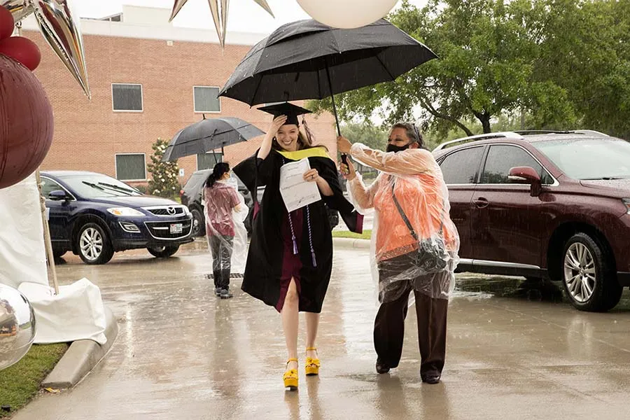 A TWU parking attendant holds an umbrella over a graduate as they walk across the sidewalk from the curb