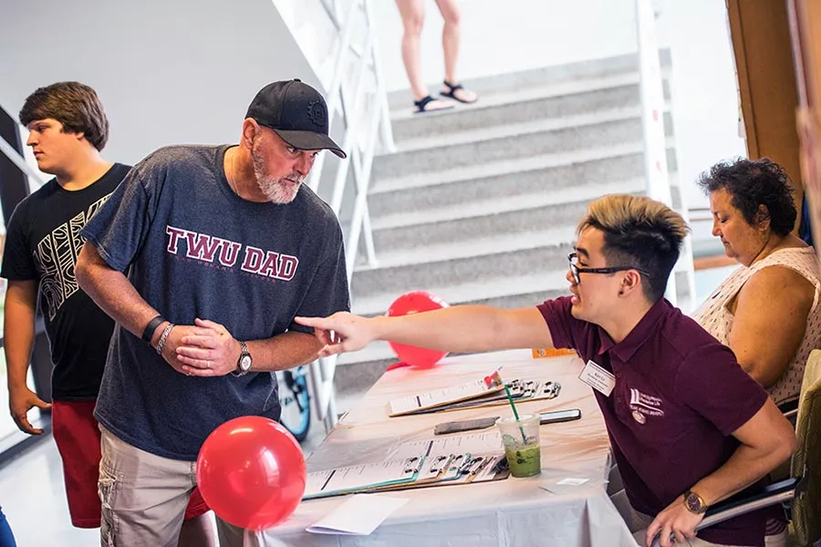 University volunteers at an information table assist a man wearing a t-shirt that says 'TWU DAD'