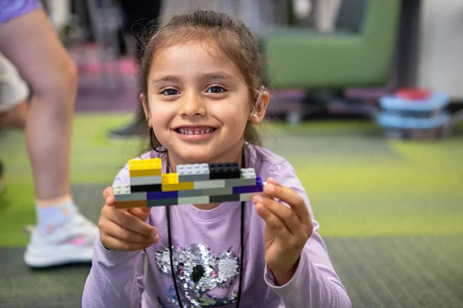 young girl in purple shirt holds up LEGOs while sitting in a carpeted room.