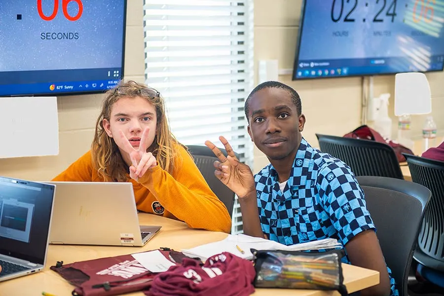 Two kids give a V symbol to the camera as they work together on a laptop