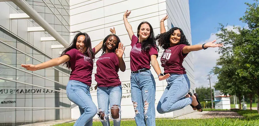 Four young women wearing TWU t-shirts leap into the air in front of the TWU Dallas campus building