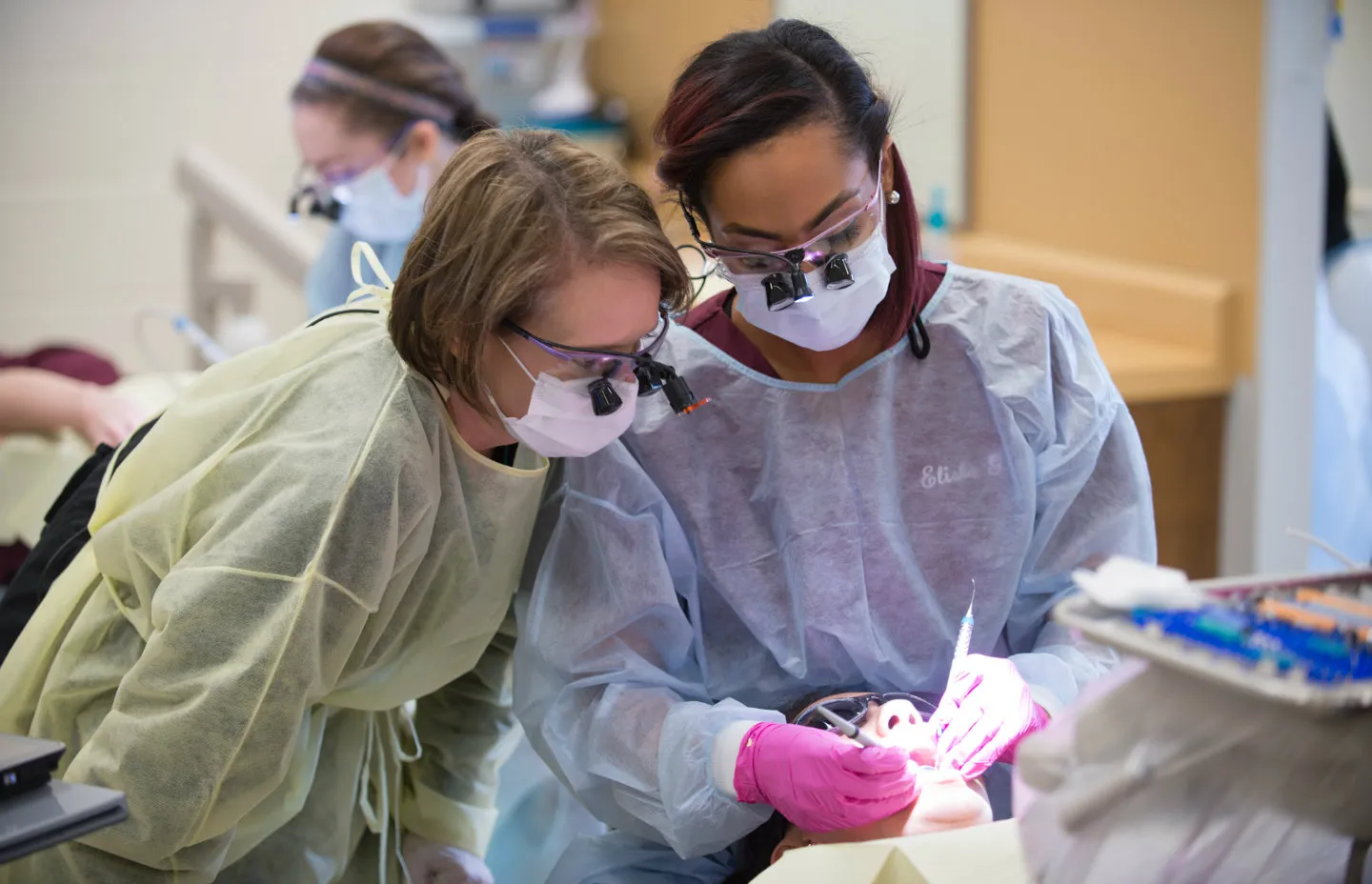 Two dental hygiene students in surgical scrubs and masks work on a patient
