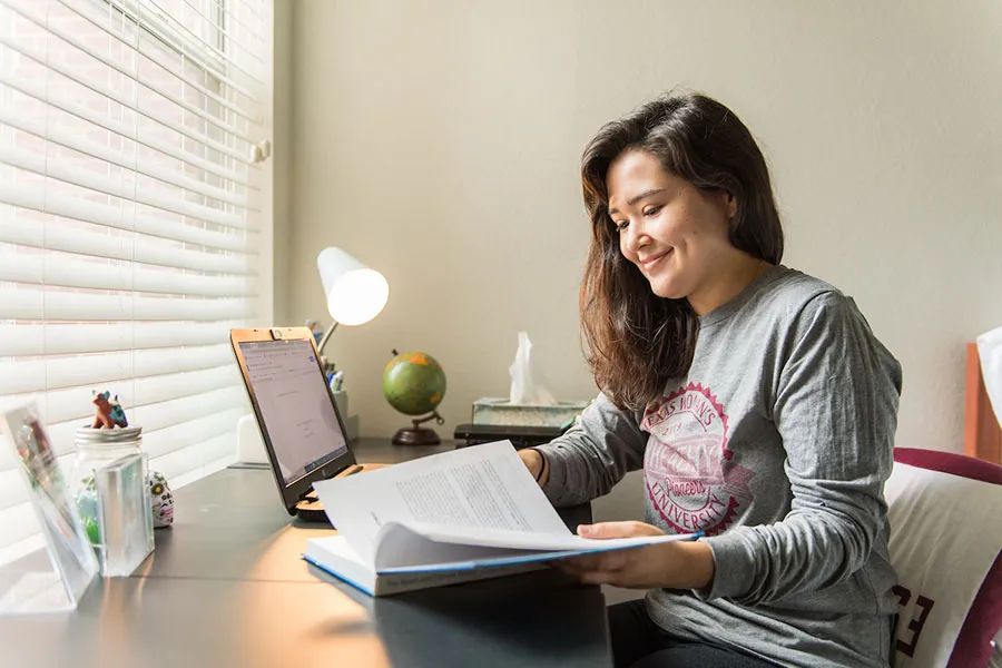 TWU student sitting at a desk reading a book next to a laptop
