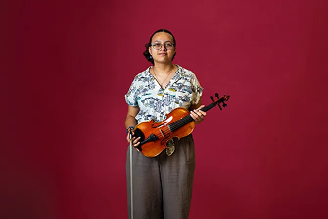 A smiling TWU student portrait on a maroon background and dressed casually and holding a violin.