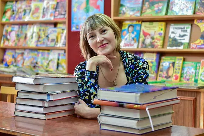 A school librarian poses for a portrait at her desk with stacks of books to her left and right on the desk.