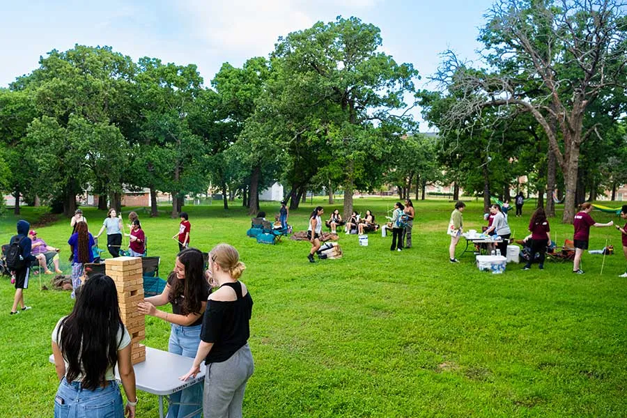 TWU students play jenga on the Lowry Woods lawn while other students mingle and socialize behind them.