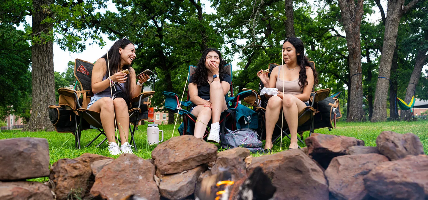 Three TWU students sit on lawn chairs around a fire while eating s'mores.