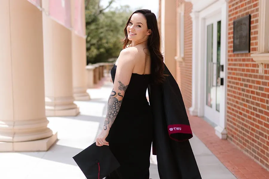 a TWU alum stands at Hubbard Hall, holding her graduation gown and cap