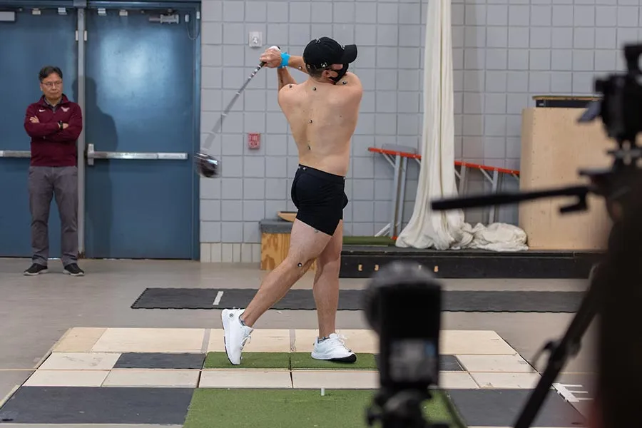 a golfer in black shorts with electrodes placeed on his body swings a golf club with Dr. Kwon behind him