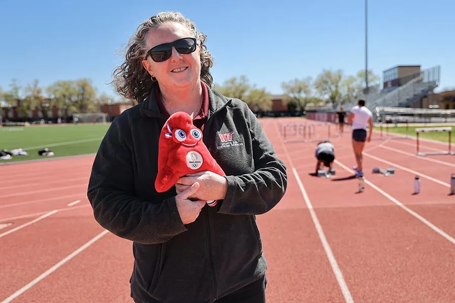 èƵ alumna standing on a track holding a small Paris Olympics stuffed mascot
