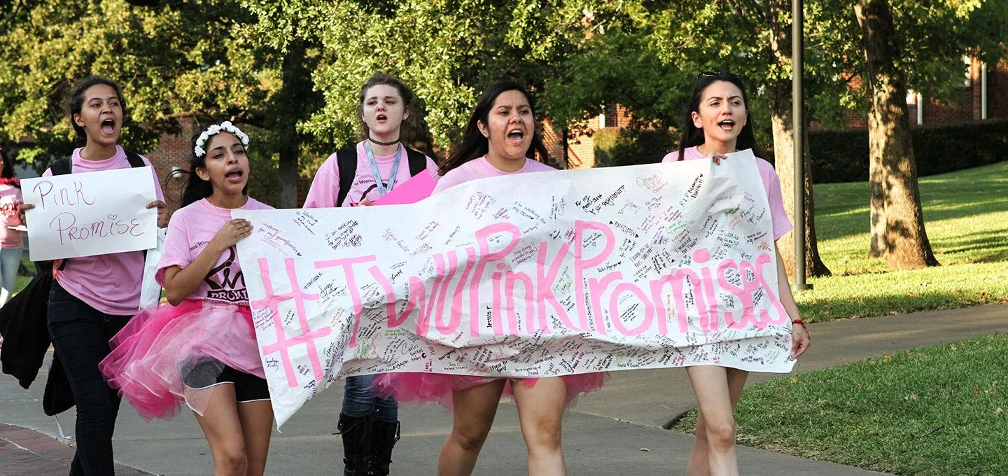 A group of students march on campus with a banner that reads #TWU Pink Promises