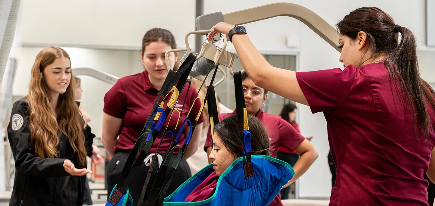 one student in maroon operates a patient lift while three students watch in the background