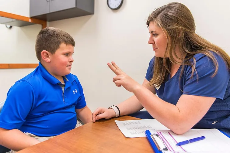 a student in blue scrubs holds up two fingers to a child sitting at a desk