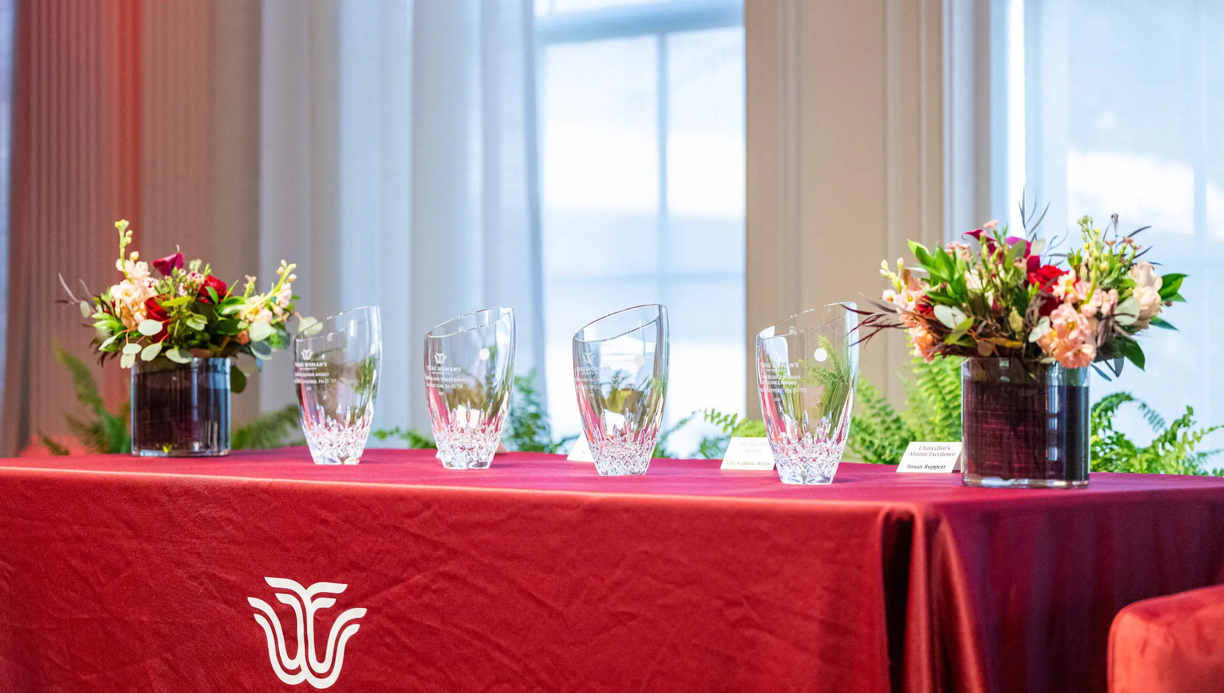 Four glass vases engraved with the award recipients' names sit on a TWU-branded tablecloth surrounded by florals.