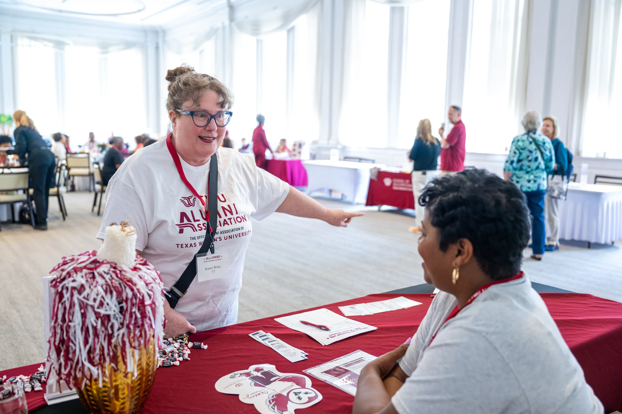 Two alumni, one sitting and hosting a table at the chapter fair and the other standing opposite her, smile and chat at the chapter fair.