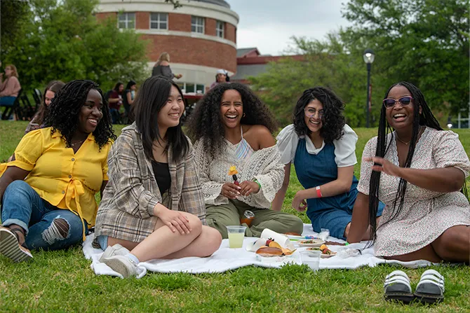 Five friends sit on the lawn near Pioneer Hall picnicing.