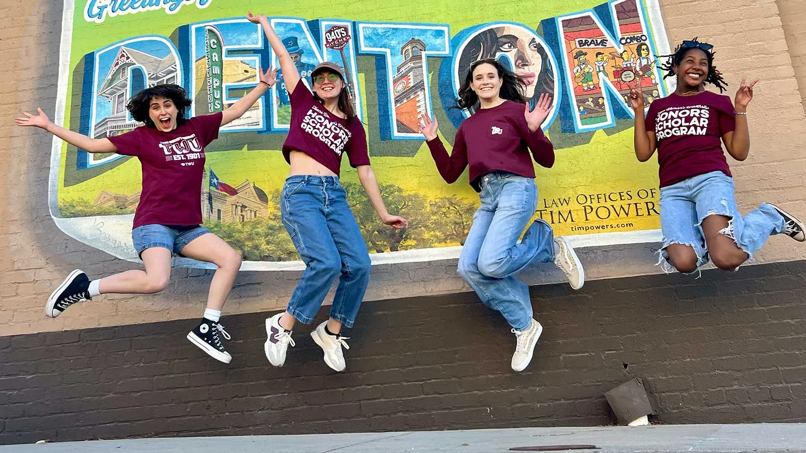 4 TWU students jumping for joy in front of a Denton mural