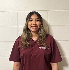 Young woman in maroon polo shirt smiles in front of a white brick background.