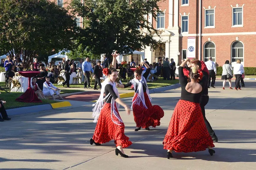 three young women dressed as flamenco dancers dance on the Pioneer Circle while a group of spectators look on from the lawn