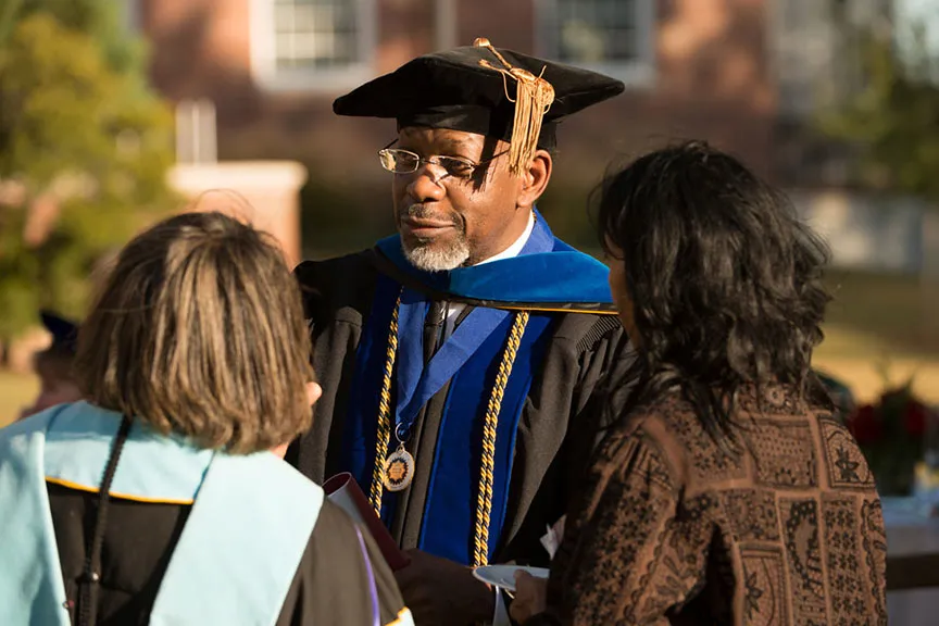 a black man in university regalia speaks with two women, one also in university regalia
