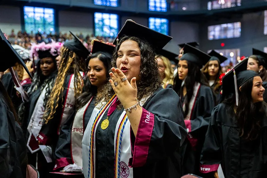 A student wearing the international scholar medallion with her cap and gown blows a kiss during Commencement