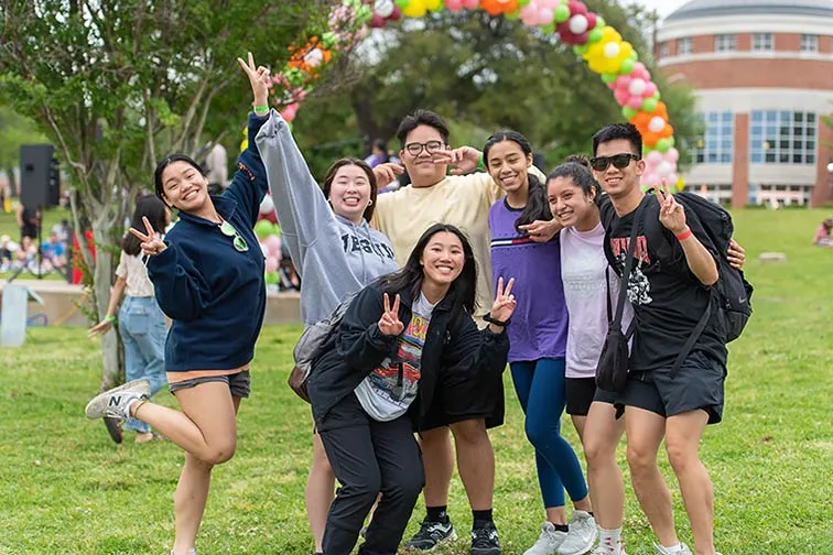 A group of people pose laughing in front of a balloon arch on the TWU Denton campus