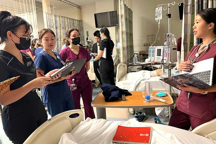 four students in scrubs holding clipboards stand around a bed in clinic setting