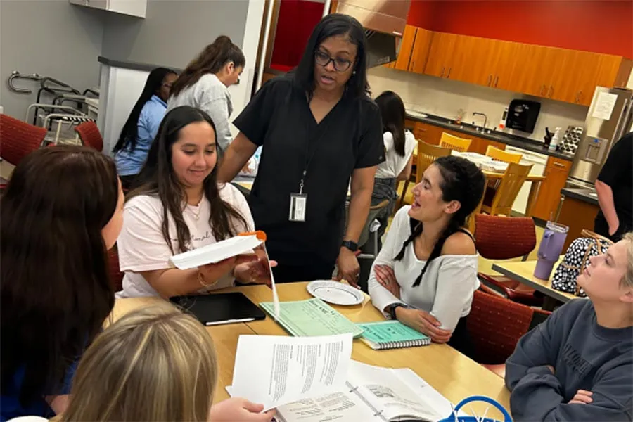 a professor talks to a group of students sitting at table in classroom