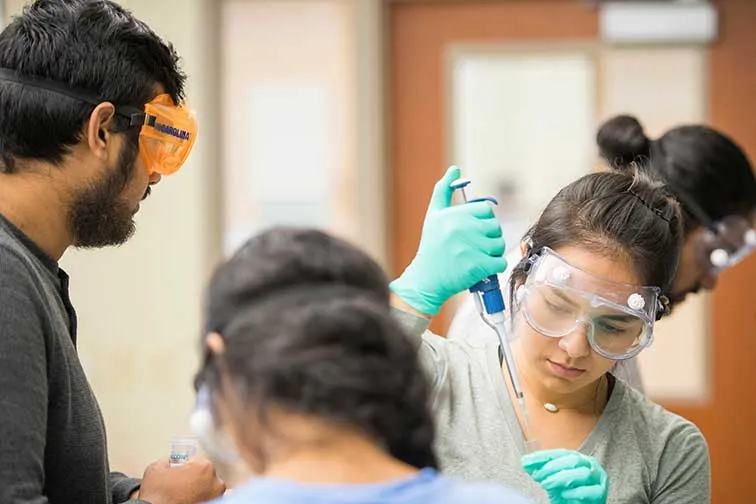 three students in goggles and scrubs work in a clinical setting