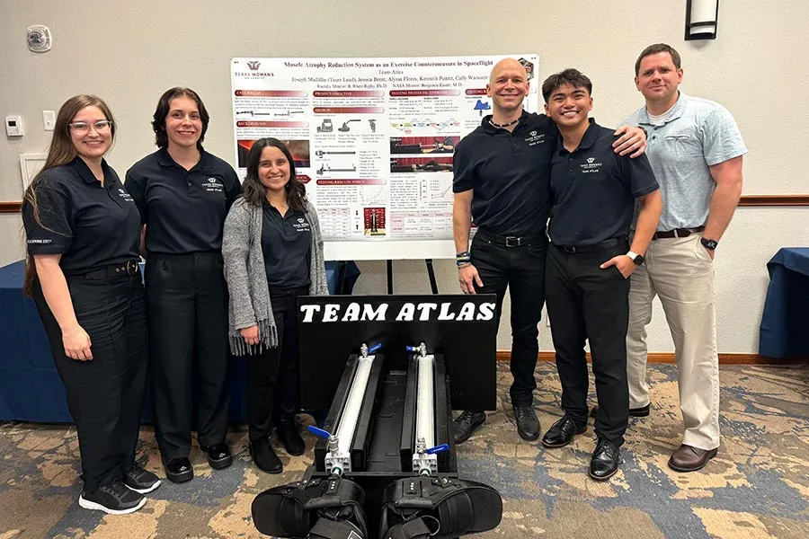 stive students dressed in black and a faculty member stand around a poster and a exercise device inside a conference room