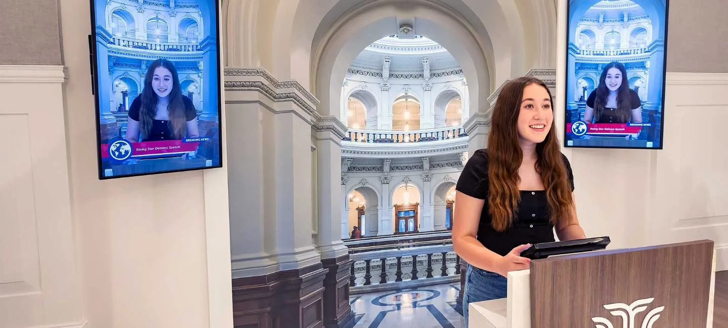 A student stands at a podium to give a speech as her image is projected onto television screens around her