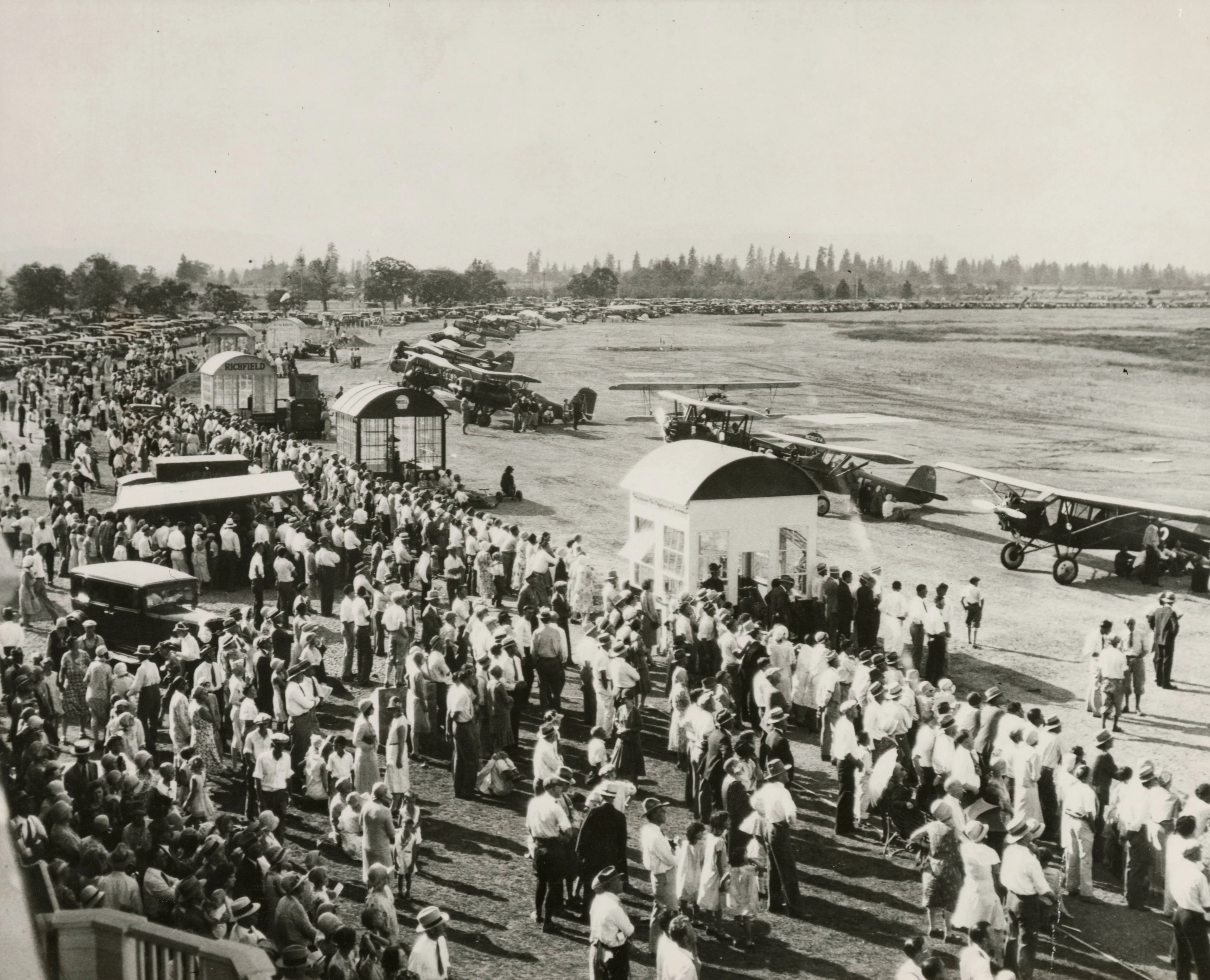 Black and white photo of a group of people on the left standing in front of a line of bi-planes on the right.
