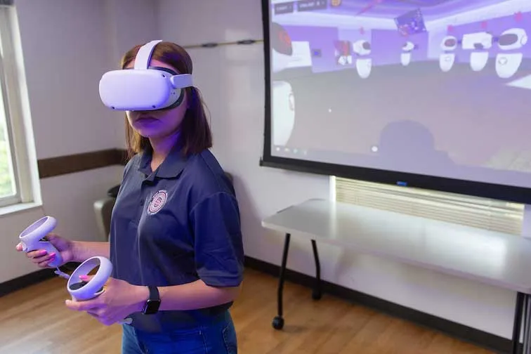 A TWU student wearing a virtual reality headset and holding hand controls interacts with a computer generated program displayed on a screen behind her.
