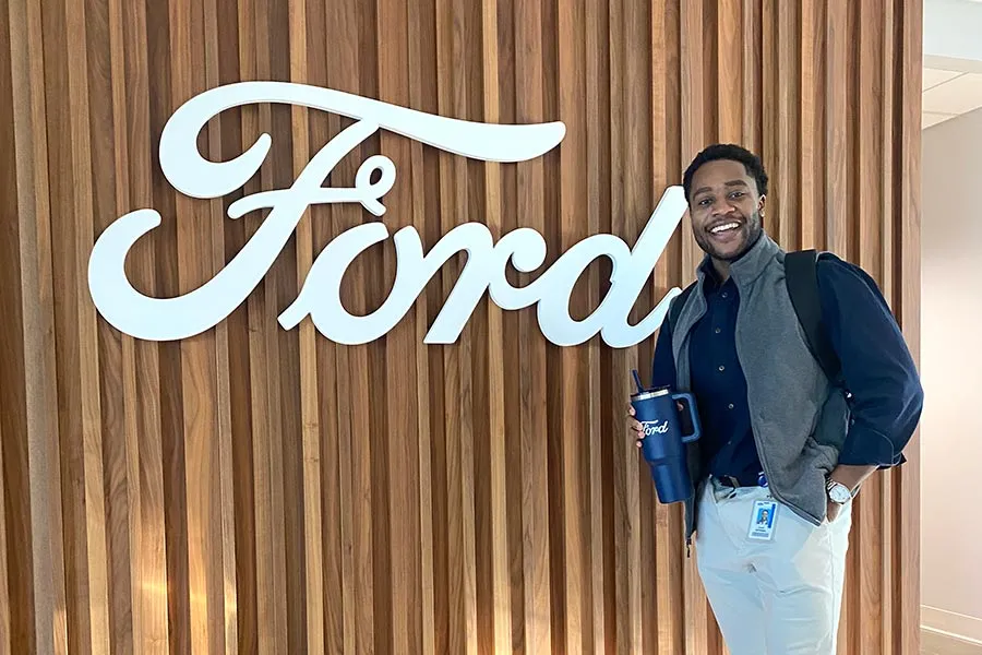 a TWU student in a gray vest stands in front of a wood panel wall with Ford  written across