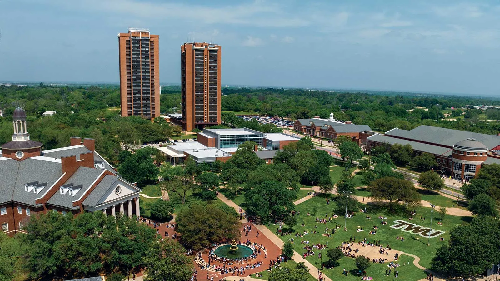 The TWU Denton campus skyline with Stark and Guinn Hall in the background and students lounging on the green lawns.