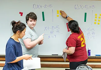 Three students work a problem on a whiteboard using rows of colored squares to visualize the process