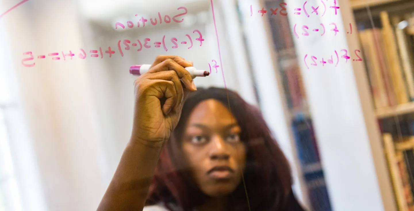 Student writing calculations on a clear board.