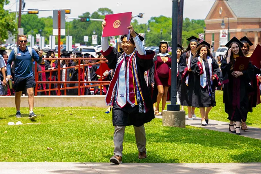 TWU McNair Scholar in cap and gown walking across the Denton campus lawn holding his diploma over his head in victory