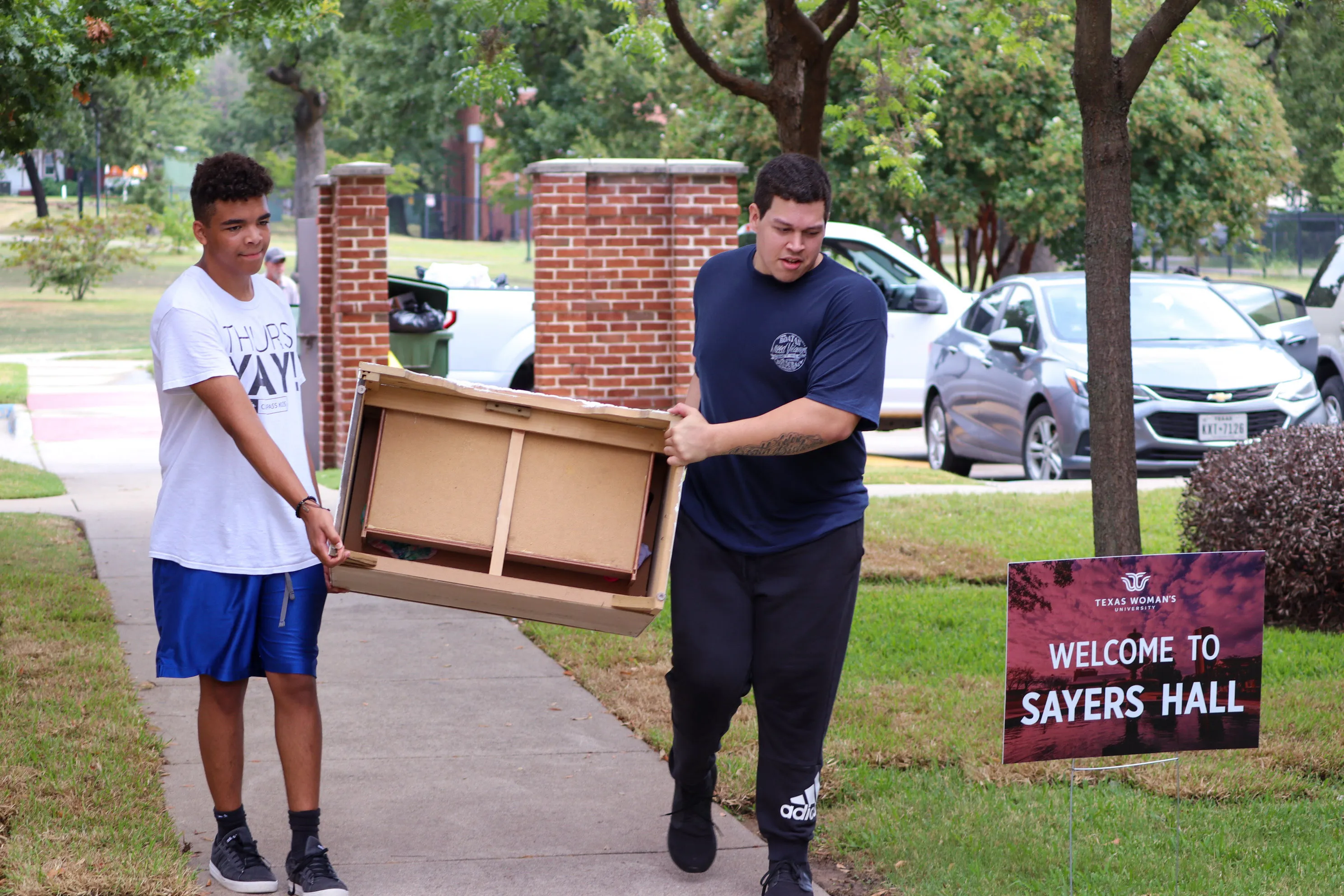 Two students transport a small dresser past a sign for Sayers Hall
