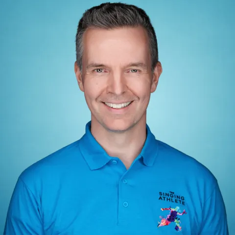 Andrew in a dark blue polo shirt against a light blue background. His shirt has a 