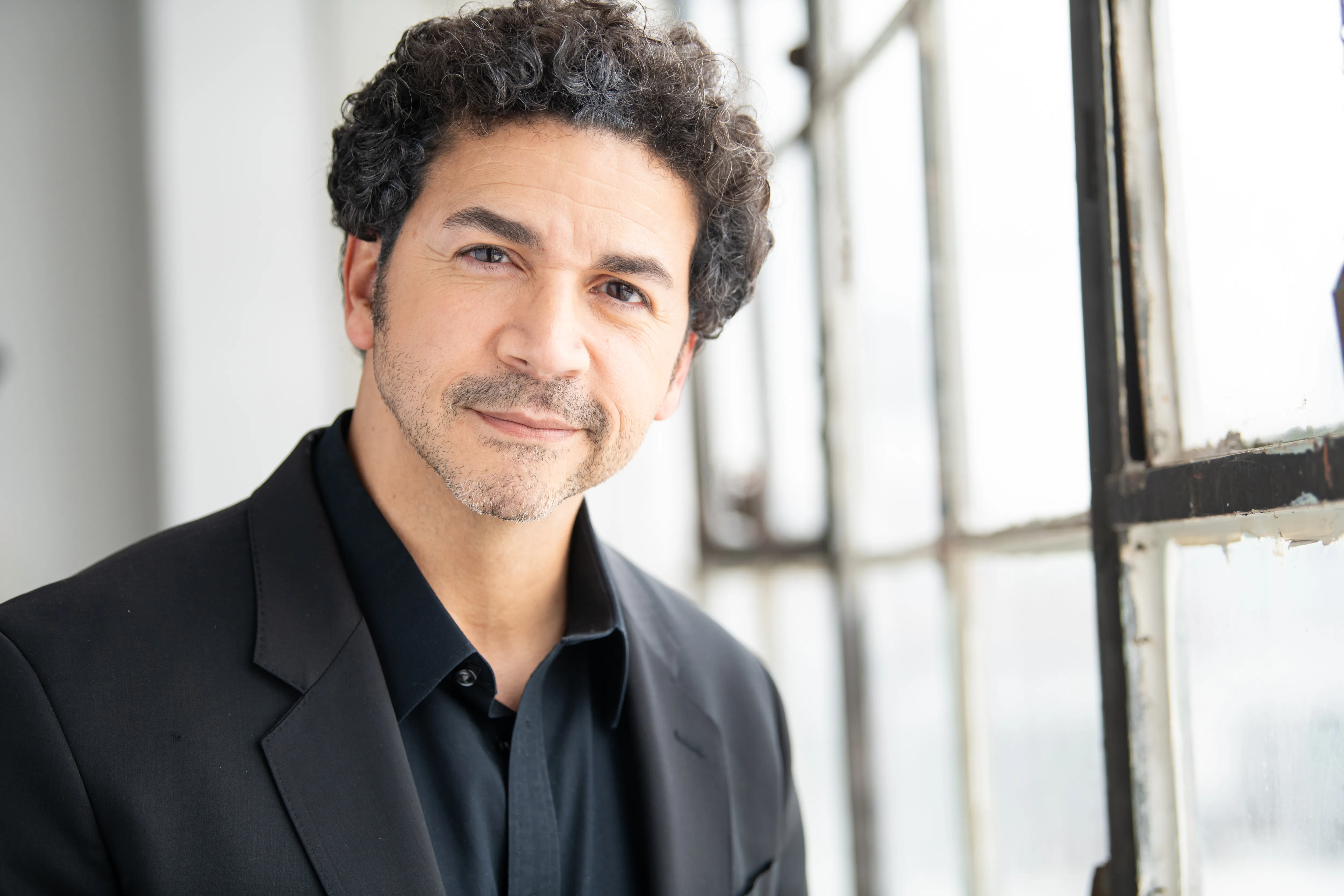 Julio in a black shirt and jacket, standing near a window