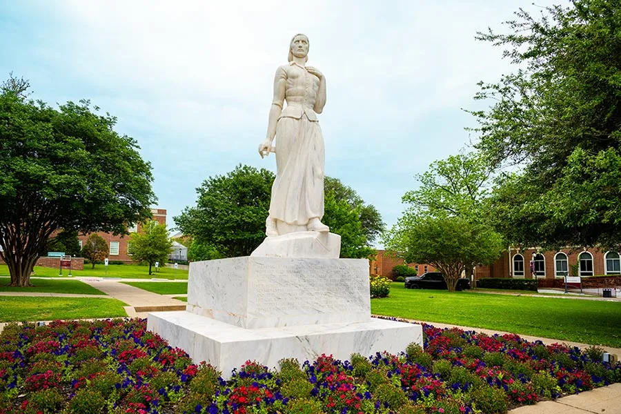 The Pioneer Woman statue surrounded by a bed of burgundy and blue flowers