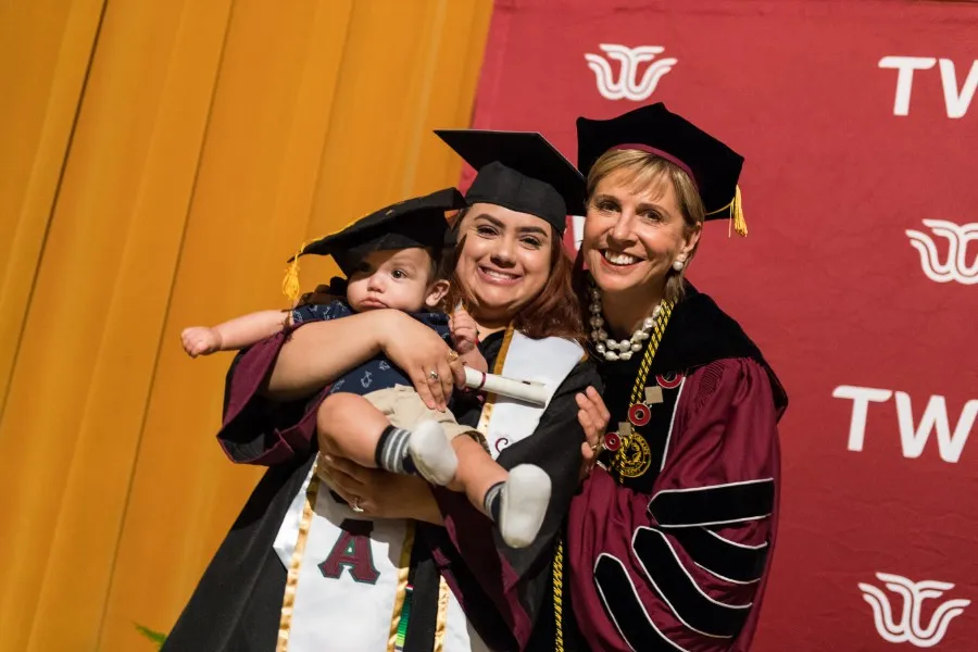 Chancellor Feyten smiles with a TWU graduate and her toddler aged son in caps and gowns.
