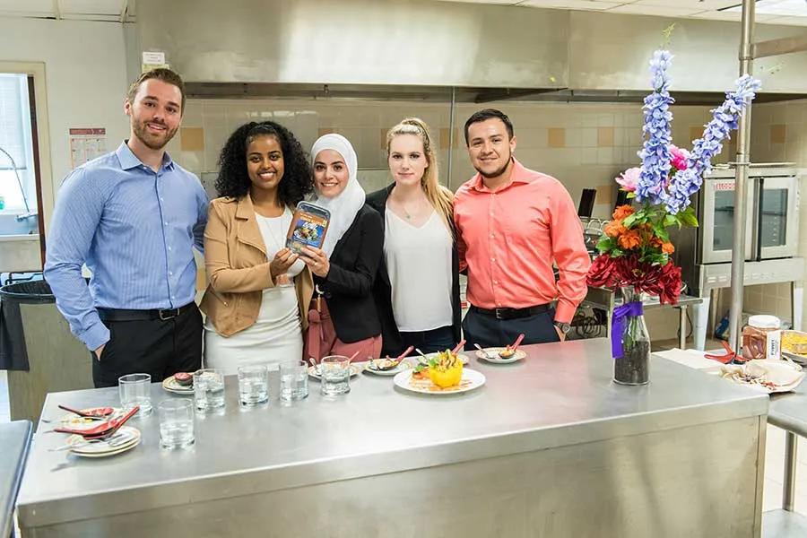 Five TWU students pose in an industrial kitchen setting with their Elote Bowls product prototype.