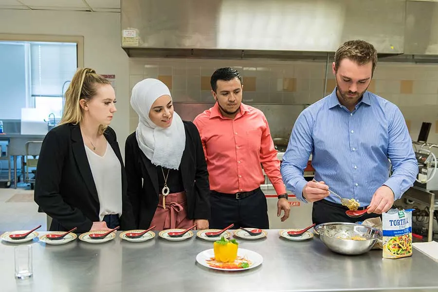 Five TWU students plate their elote food product in an industrial kitchen setting.	