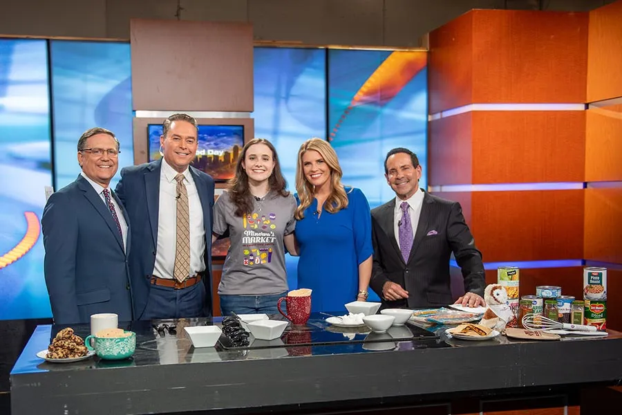 nutrition student in gray shirt stands behind news desk with four hosts from the Good Day morning show