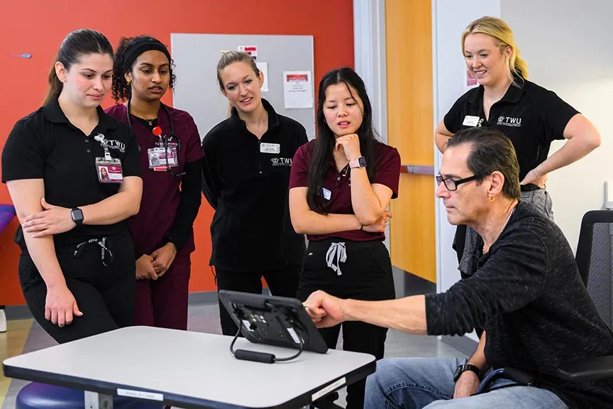 five Occupational Therapy students stand around a desk while a patient sits behind a computer